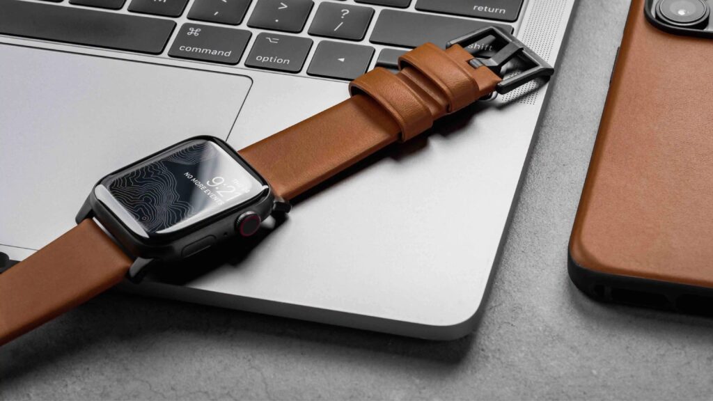 Apple Watch with a brown leather band resting on a laptop beside a matching leather phone case