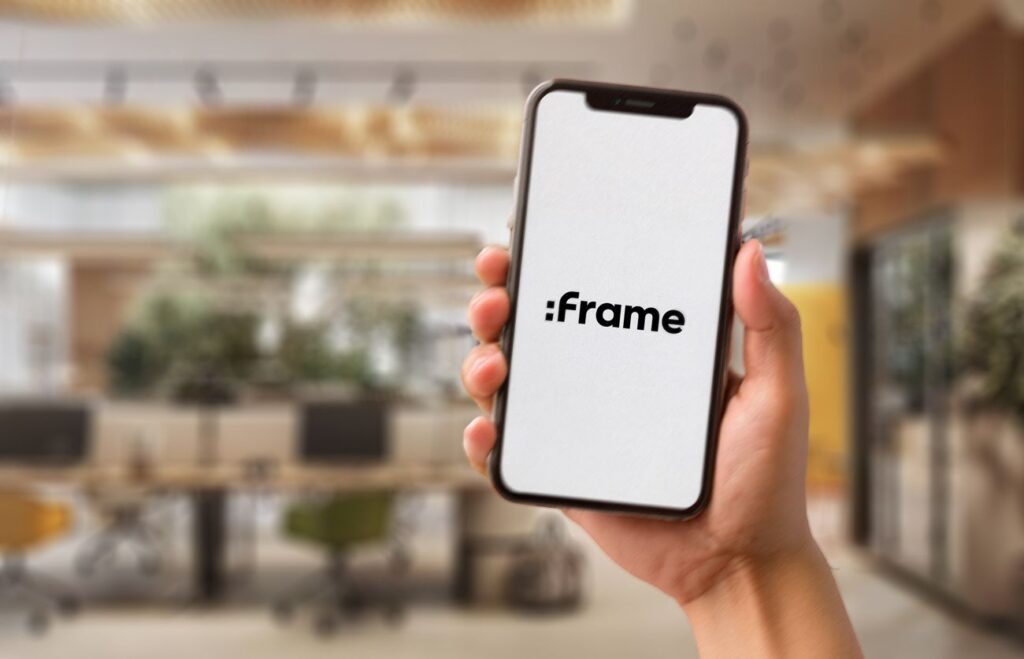 Hand holding smartphone displaying iFrame logo against blurred indoor background.