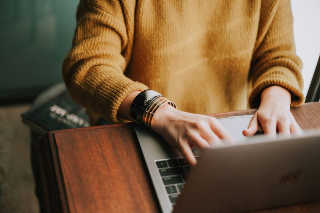 Person in mustard sweater types on laptop while holding a leather-bound notebook at a wooden desk.