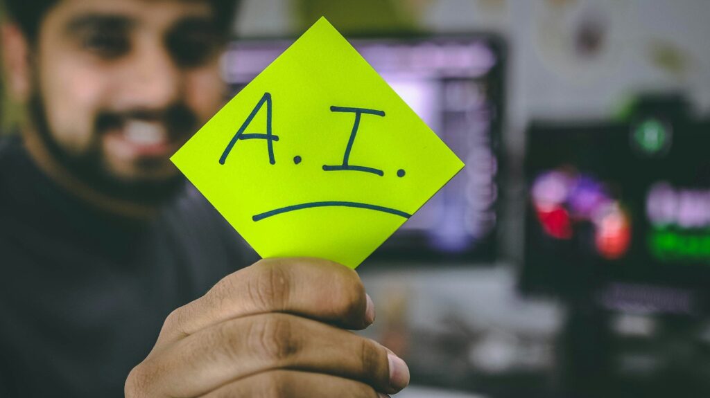Person holding a bright green sticky note with the text "A.I." written on it in front of a blurred computer workspace