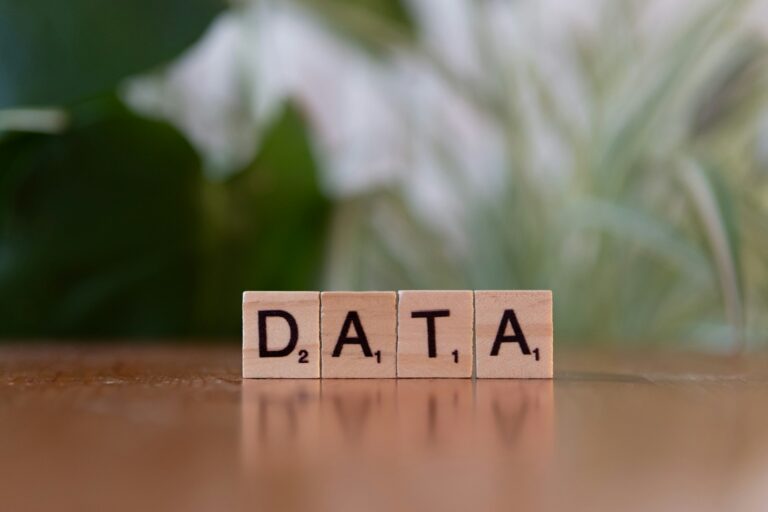 Wooden Scrabble tiles spelling DATA on a reflective surface with blurred green foliage behind.