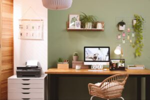 How to Budget for Remote Work: From Home Office Design to Tech Investments A home office with sage green walls features a wooden desk, wicker chair, dual monitors, and organized wall shelves