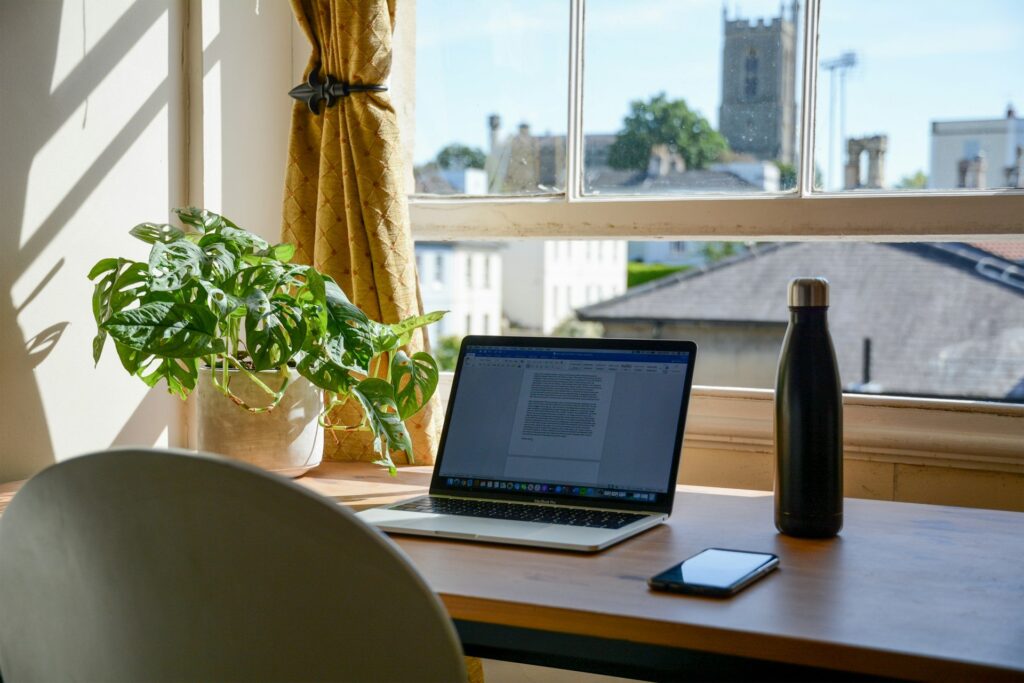 Sunlit desk with laptop, potted plant, and water bottle overlooking a historic stone church tower.