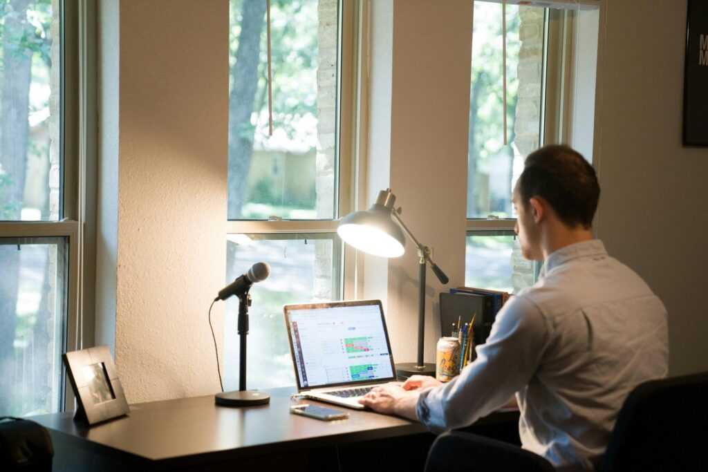 Man working at desk with laptop near large windows overlooking trees, with microphone and desk lamp.
