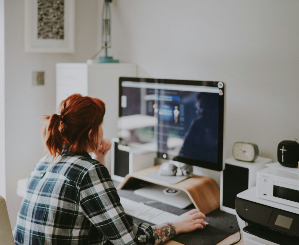 Person with red hair in plaid shirt works at a minimalist desk with desktop monitor in a bright, white home office.