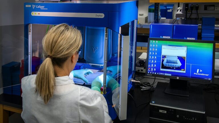 Laboratory technician in protective gloves operates a blue Caliper Zephyr automated liquid handler in a research facility.