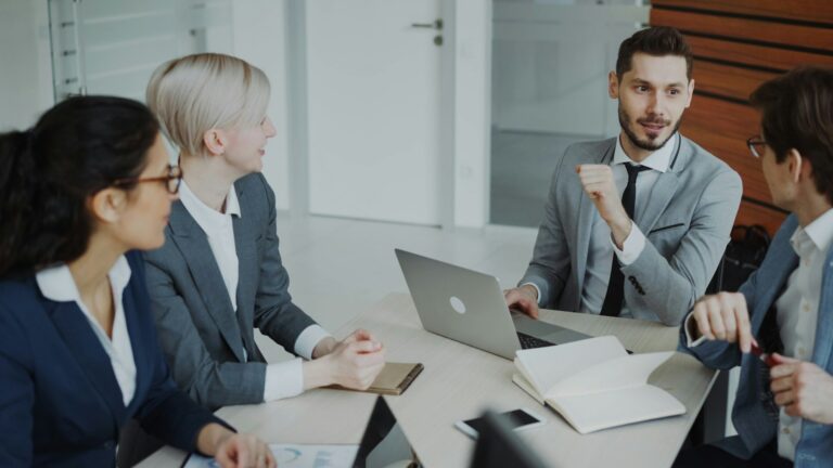Four business professionals in suits discuss strategy around a conference table with a laptop and documents.