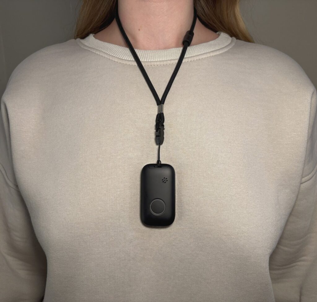Black mobile alert device with a round button hanging from a black lanyard over a cream crew-neck sweatshirt