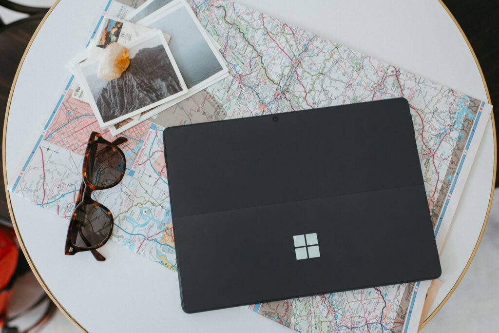 Overhead view of a white round table with a laptop, printed maps, sunglasses, and travel photos arranged for trip planning.