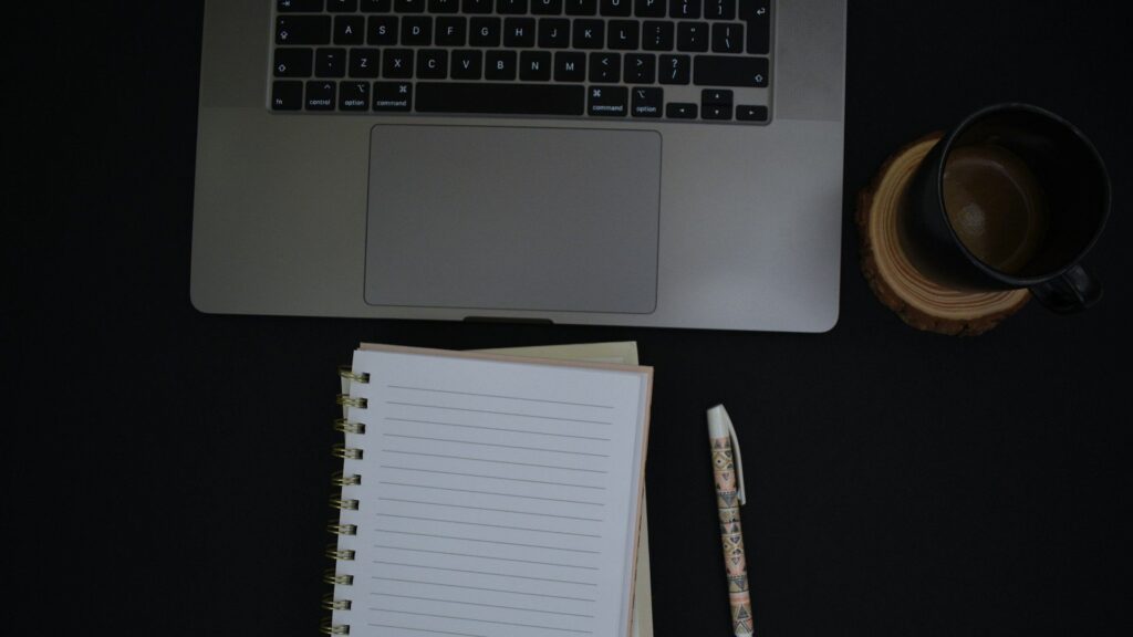 Overhead view of a workspace with a silver laptop, spiral notebook, decorative pen, and coffee mug on a dark surface.