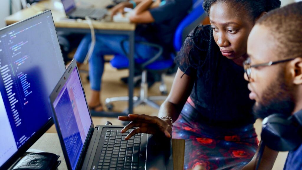 Two people examine code on a laptop screen during a collaborative programming session in a classroom setting.